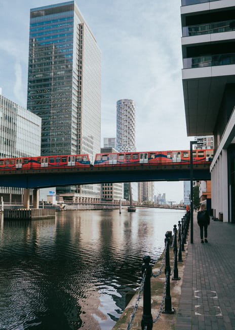A cityscape along a river featuring a red train crossing a bridge overhead, surrounded by modern high-rise buildings with glass facades reflecting the sky. The river below has gentle ripples and is bo