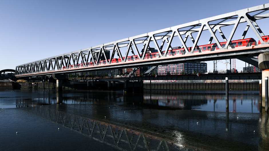 A cityscape along a river featuring a red train crossing a bridge overhead, surrounded by modern high-rise buildings with glass facades reflecting the sky. The river below has gentle ripples and is bo